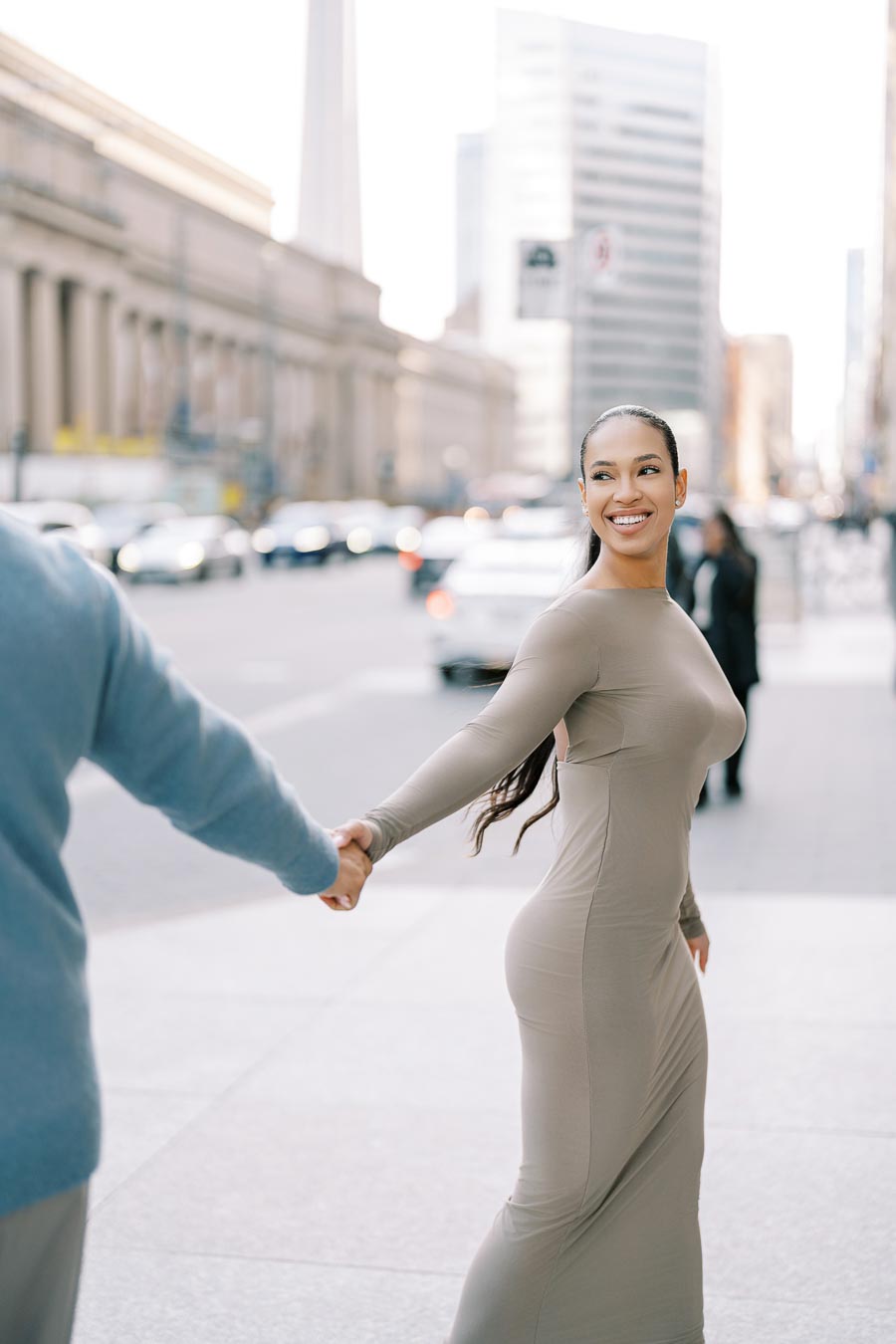 A woman in a form-fitting dress smiles as she holds hands with someone on a bustling city street, with tall buildings in the background.