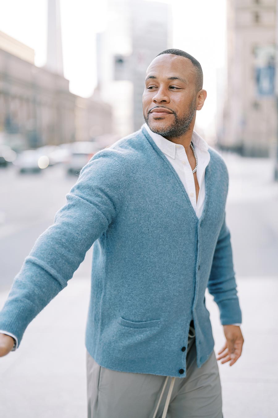 A man in a blue cardigan and white shirt walks confidently in a city environment.