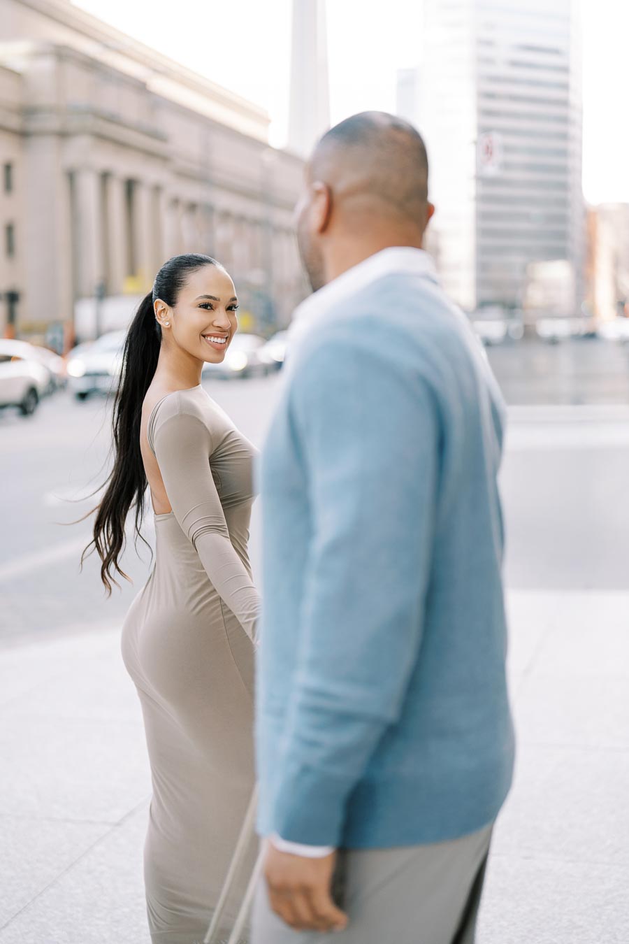 A woman in a beige dress smiles at a man in a blue sweater on a city street, with urban buildings in the background.
