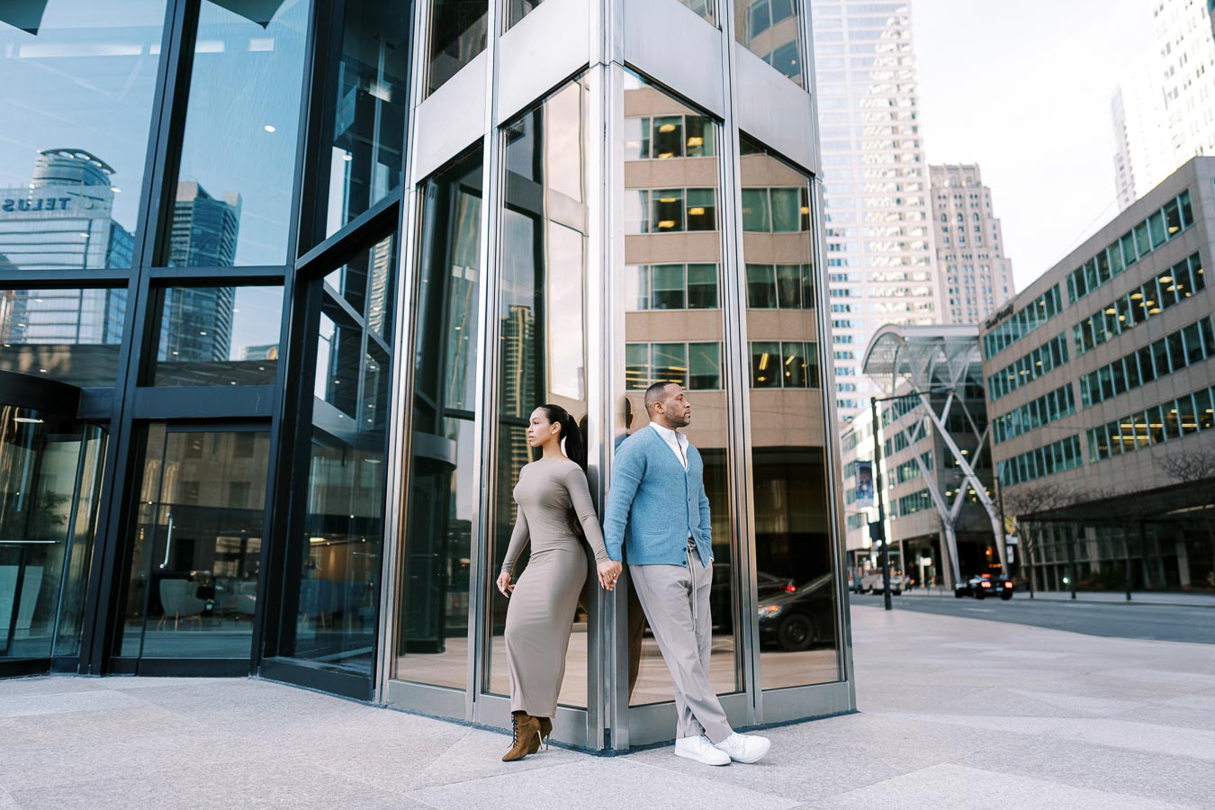 A couple stands back-to-back outside a modern glass building in a cityscape, showcasing urban architecture and style.