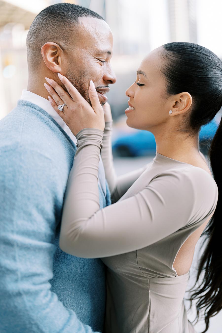 A couple shares a tender moment, embracing and smiling at each other outdoors. The woman is wearing a ring and has her hands gently on the man's face.