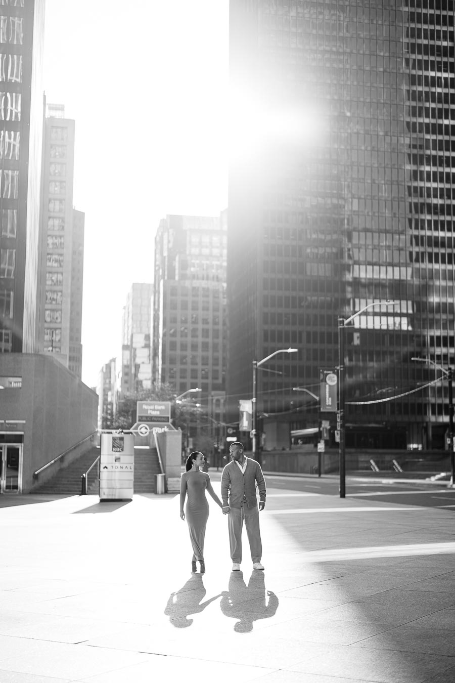 A couple walks hand in hand in a cityscape, bathed in sunlight and surrounded by towering buildings.