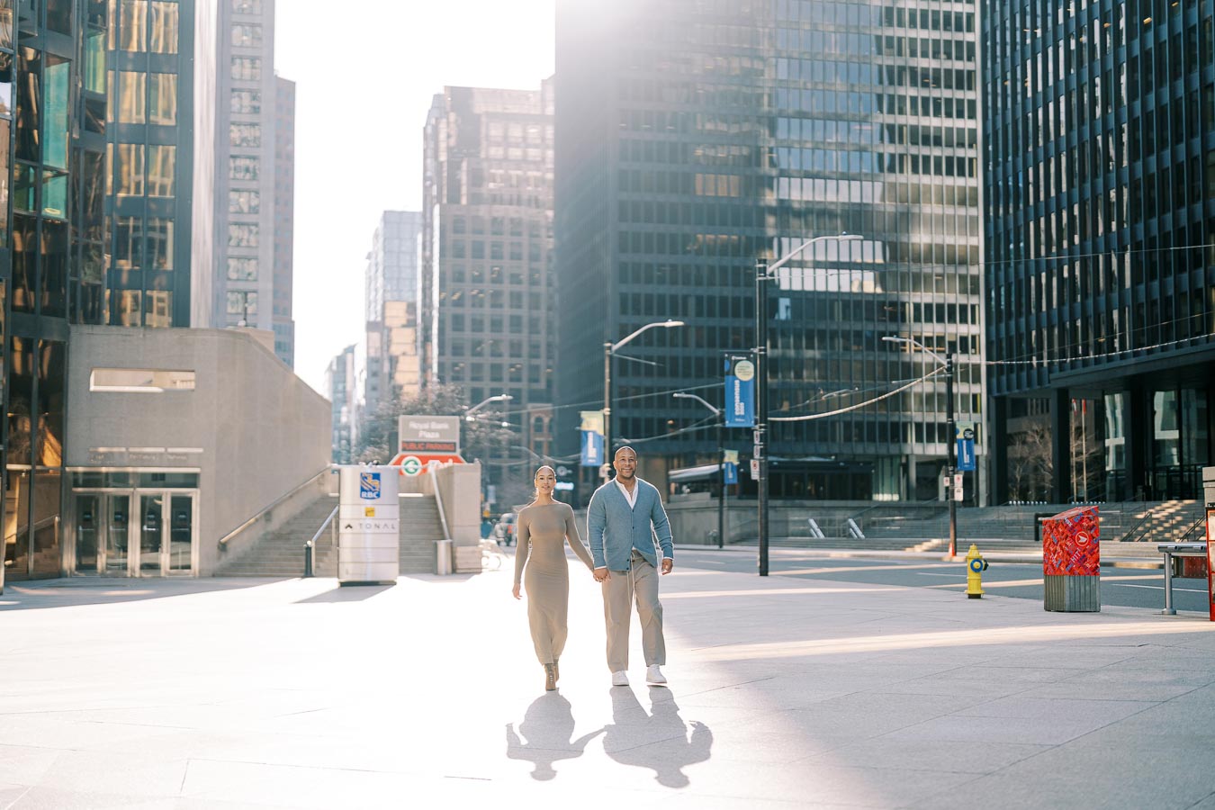 A couple walks hand in hand through a modern cityscape, surrounded by tall buildings and bathed in sunlight.