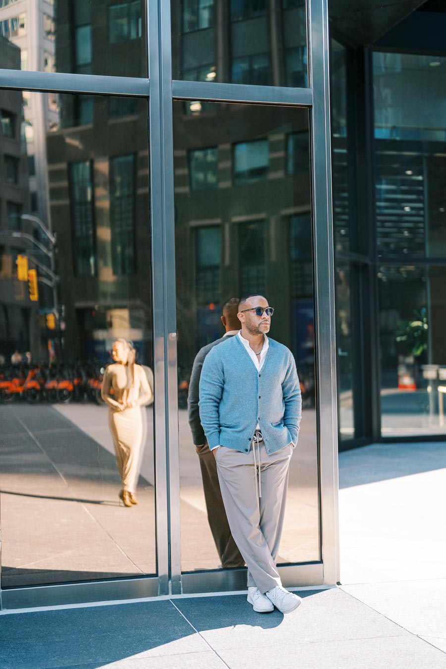 A stylish man leans against a glass building, with a woman's reflection visible in the glass.