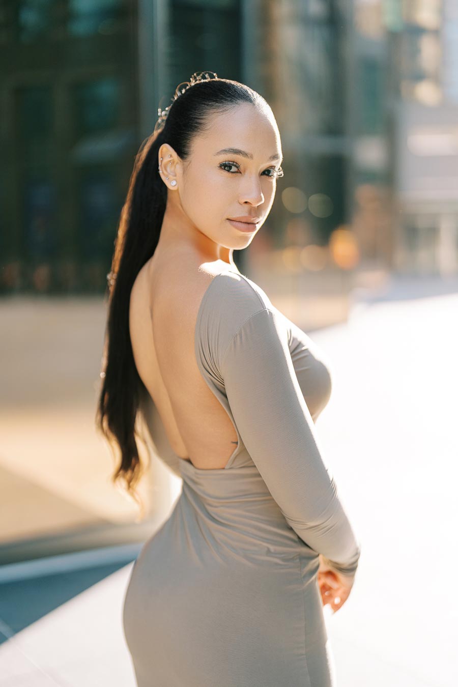 A woman poses confidently in a backless dress, with a cityscape softly blurred in the background, highlighting her elegance and style.