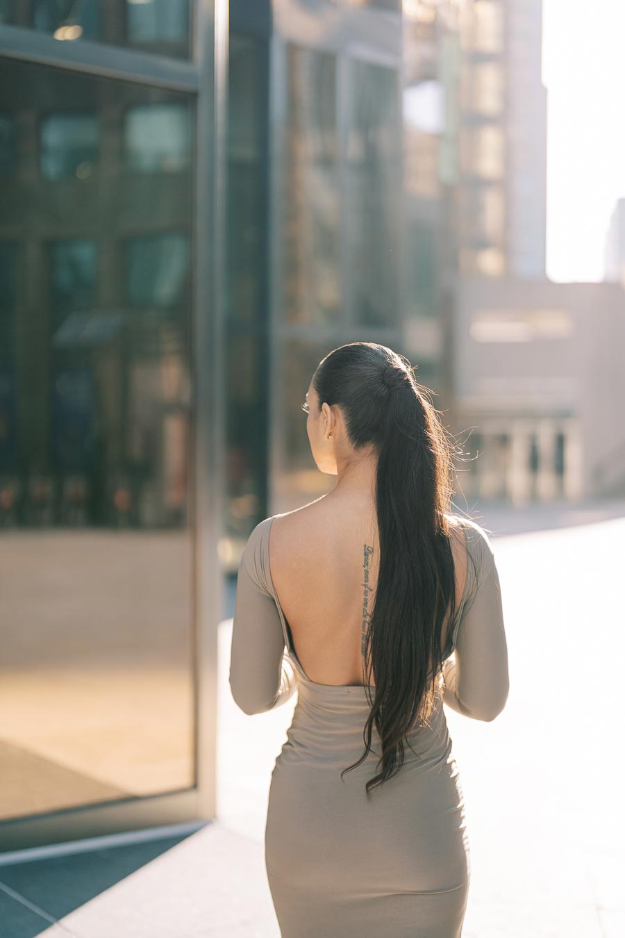 A woman with long hair in a backless dress stands in an urban environment with modern glass buildings.