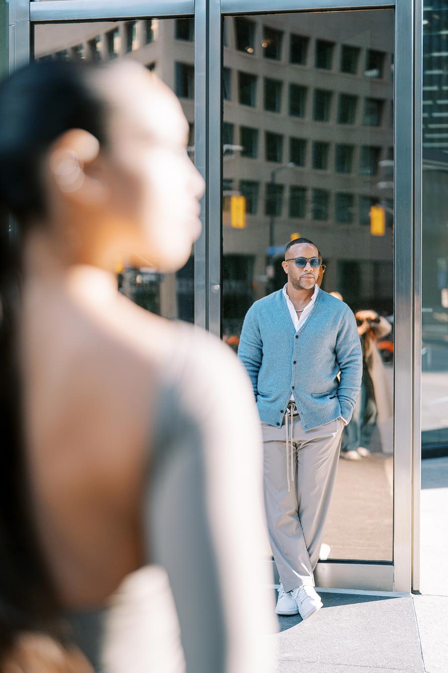 A stylish man in a blue cardigan and sunglasses leans against a glass building, with a blurred figure in the foreground, creating a depth of field effect.