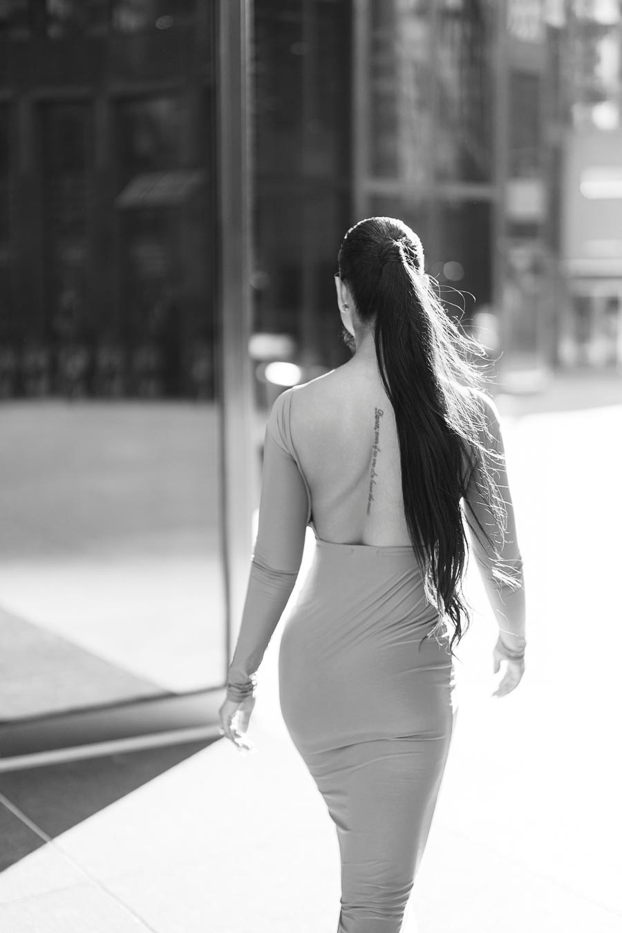 A woman with long hair walks confidently in a backless dress, captured in a black and white urban setting.