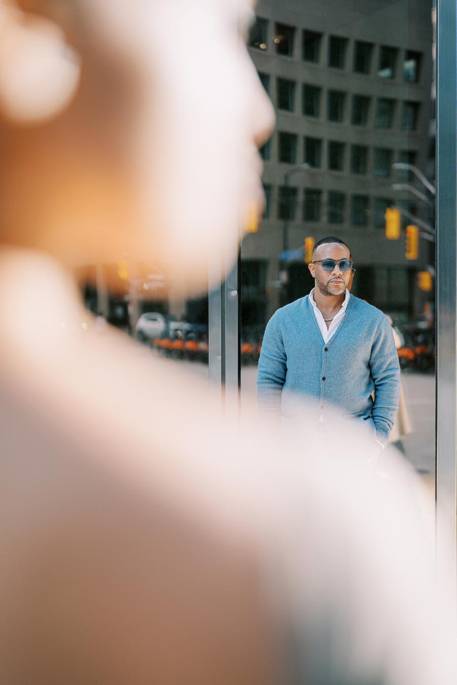 A man wearing sunglasses and a blue cardigan is reflected in a glass window, with a blurred figure in the foreground, creating an artistic composition.