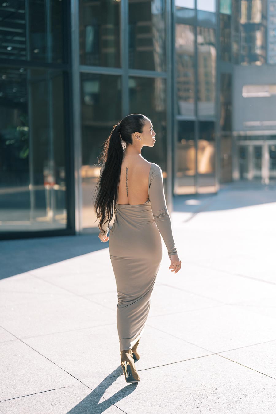 A woman in a long, backless beige dress walks confidently in an urban environment, surrounded by modern glass buildings.