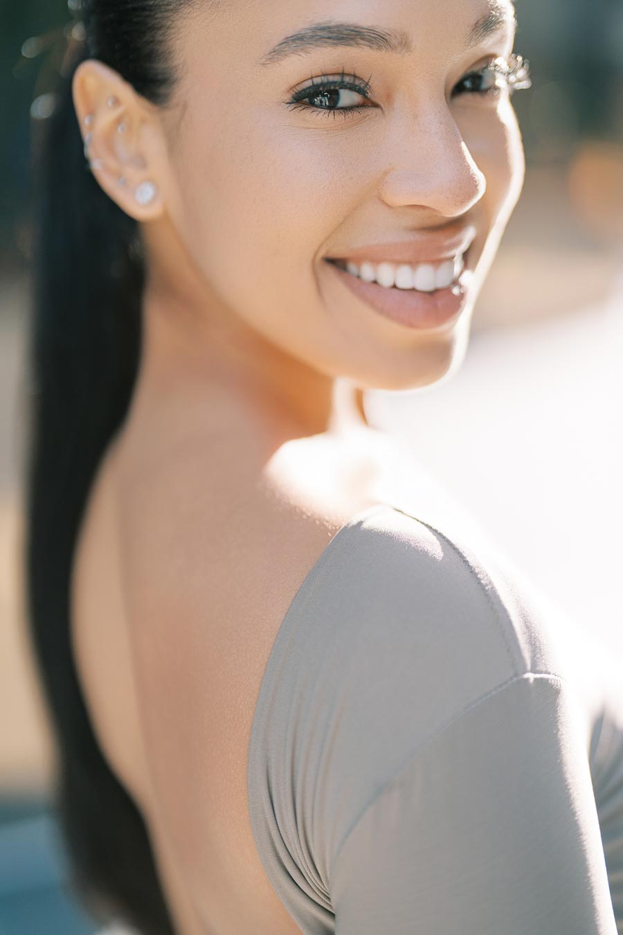 A close-up portrait of a smiling woman with long dark hair and multiple ear piercings, wearing a gray top.