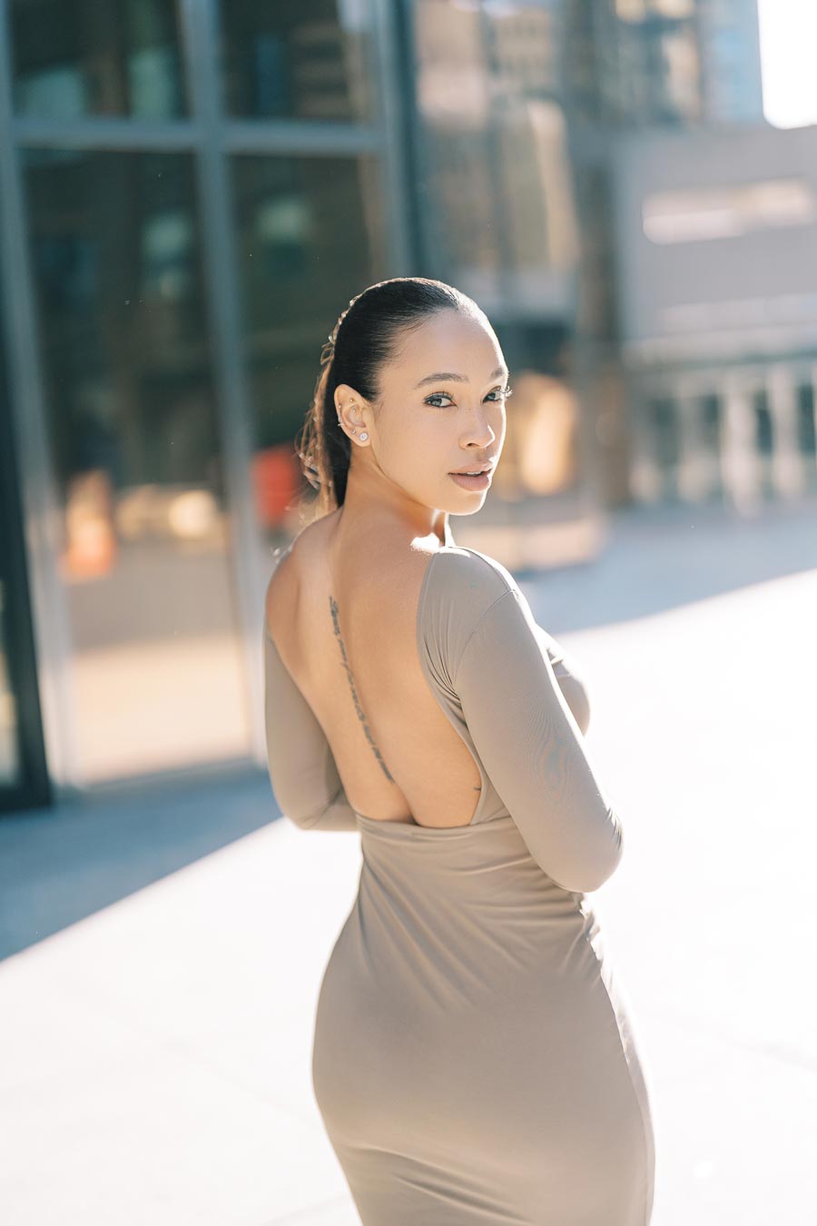A woman wearing a backless beige dress poses confidently in an urban environment, with modern buildings in the background.