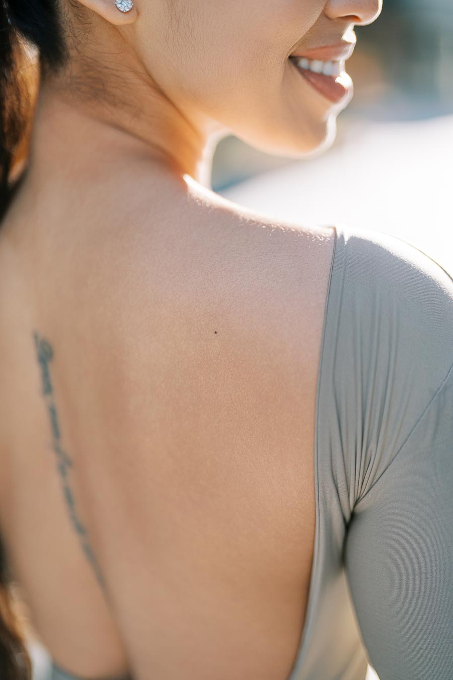 A close-up image of a woman's back, showcasing a tattoo along her spine. She is wearing a gray top with an open back, and her face is partially visible with a smile. The lighting is soft and natural, highlighting her skin tone.