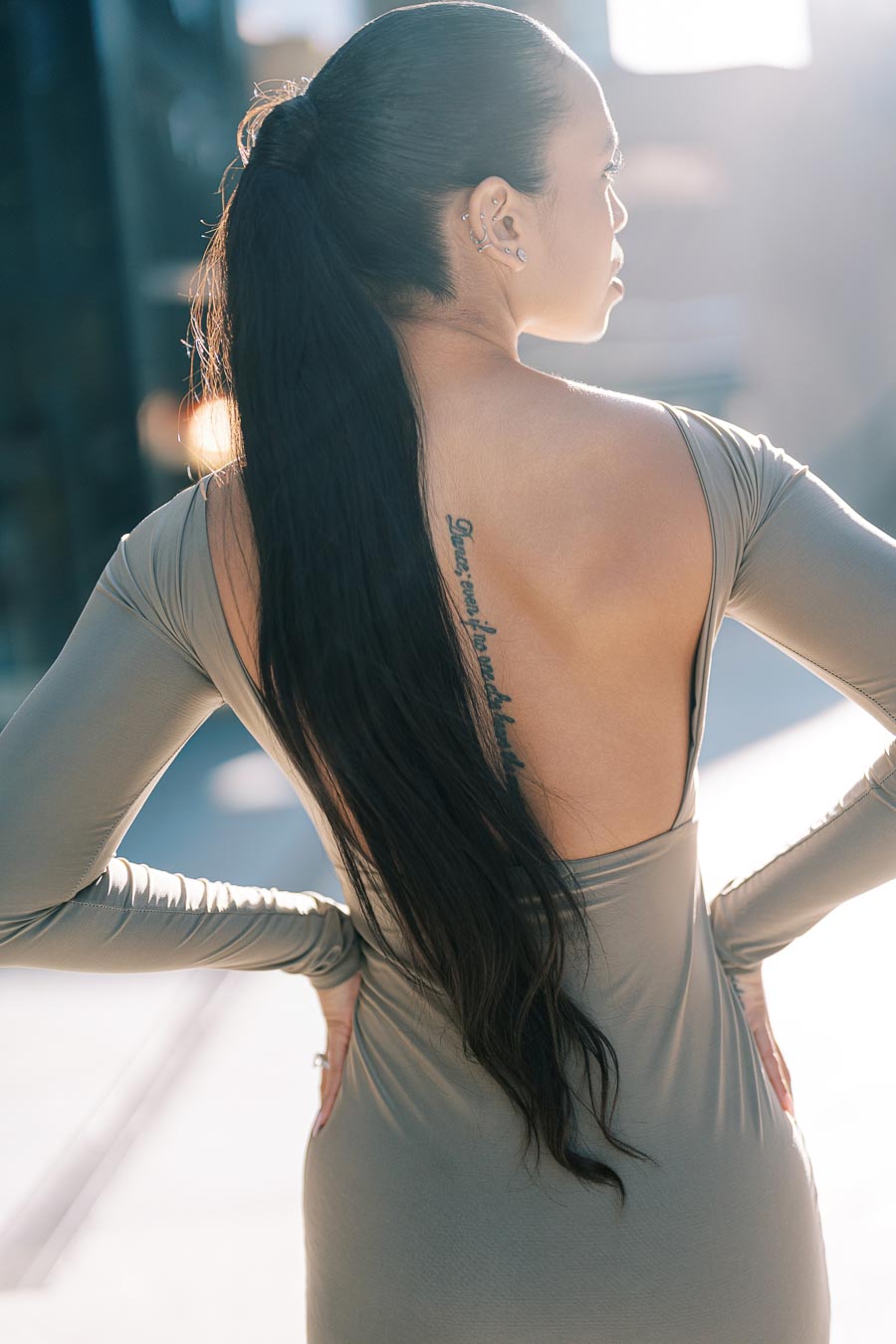 A woman with a long ponytail and a back tattoo poses in a backless dress, illuminated by soft sunlight.
