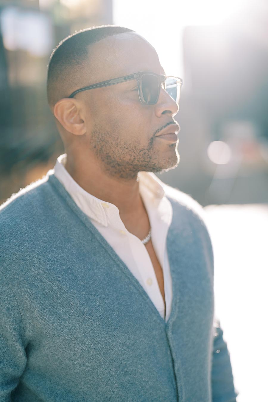 A man in sunglasses and a blue sweater stands outdoors, bathed in sunlight.