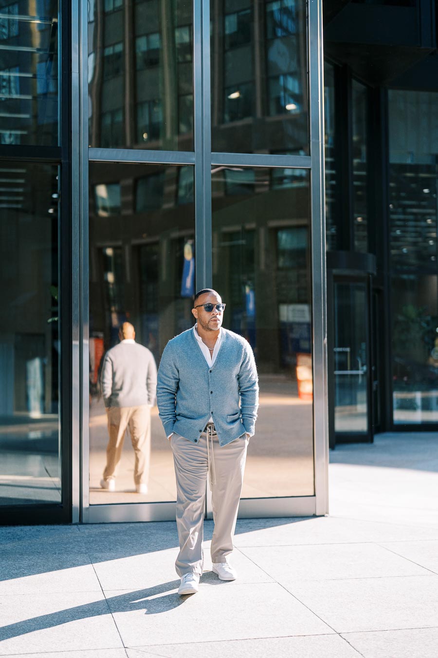 A stylishly dressed man stands confidently in front of a modern glass building, with his reflection visible in the window.