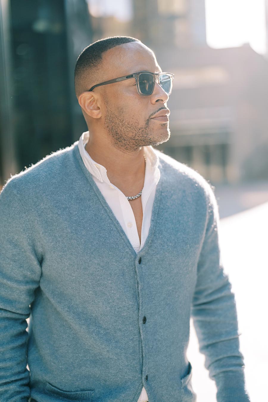 A man in sunglasses and a blue cardigan enjoys a sunny day outdoors.