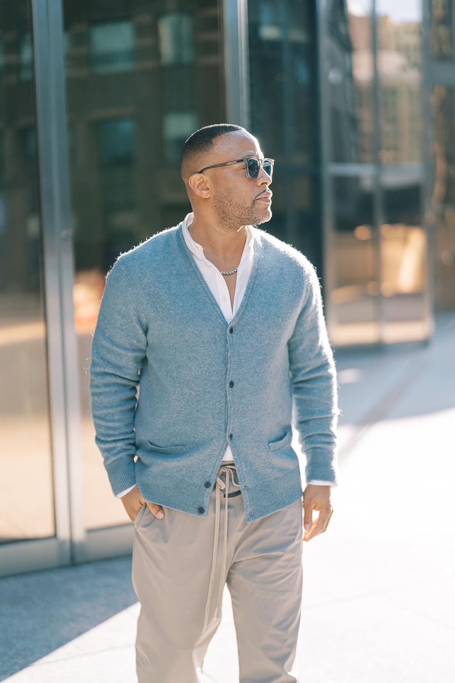 A stylish man in sunglasses and a light blue cardigan walks confidently in an urban environment.