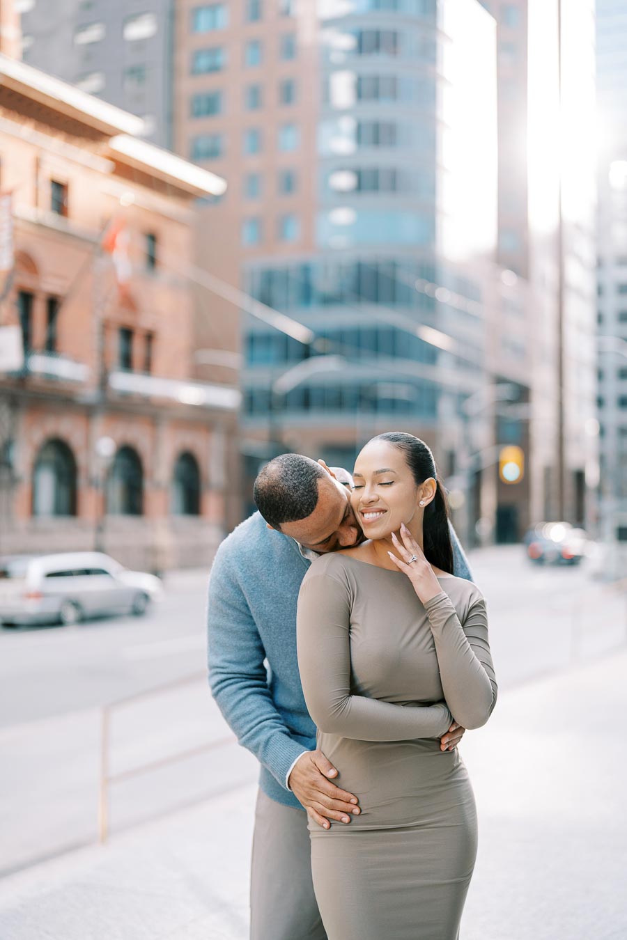 Groom-to-be kissing fiancée’s shoulder while she smiles during engagement session on downtown Toronto street