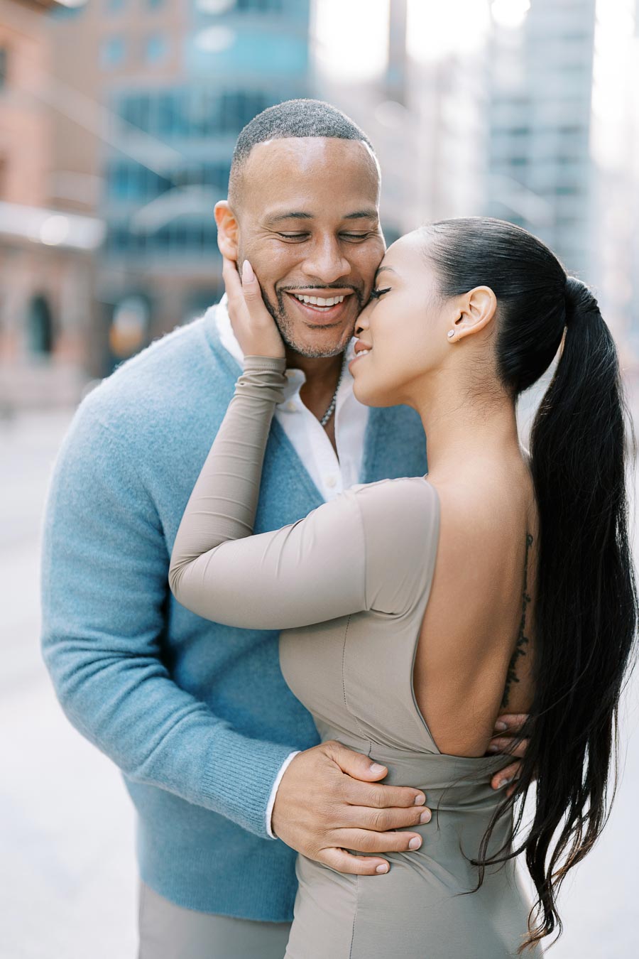 A couple shares a tender moment in an urban setting, with the man wearing a blue sweater and the woman in a beige dress.