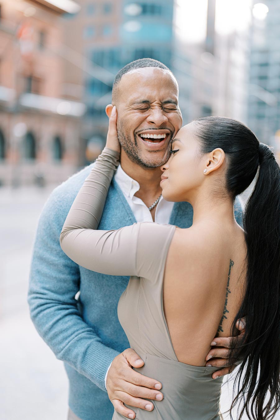 A joyful couple shares an intimate moment in a city setting, with the man laughing and the woman gently holding his face.