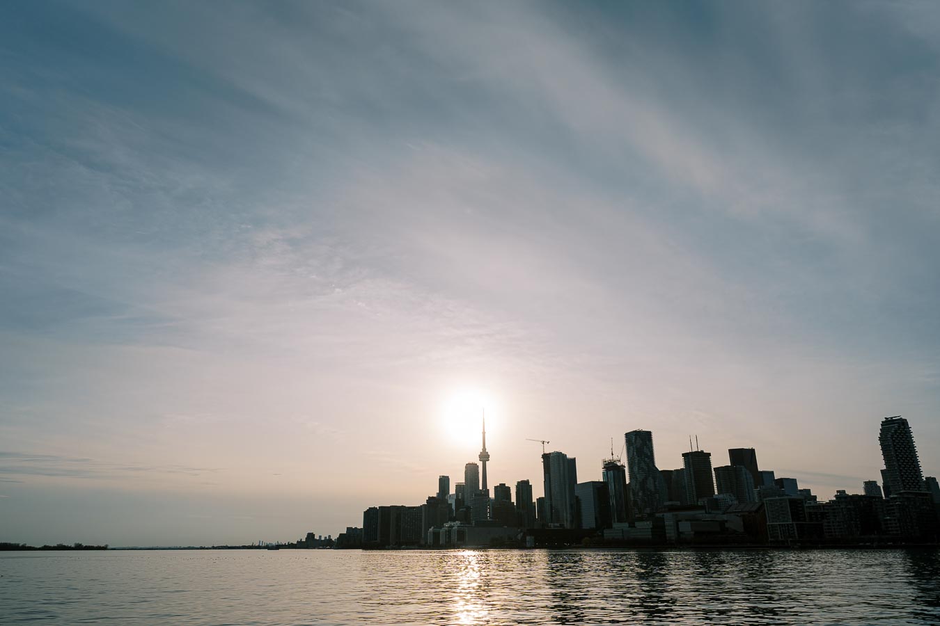 The Toronto skyline is silhouetted against a setting sun, with the iconic CN Tower prominently visible. The scene is viewed from across the water, creating a serene and picturesque cityscape.