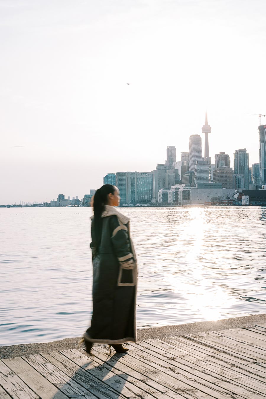 A person stands on a wooden dock overlooking the water, with the Toronto skyline and CN Tower visible in the background during sunset.