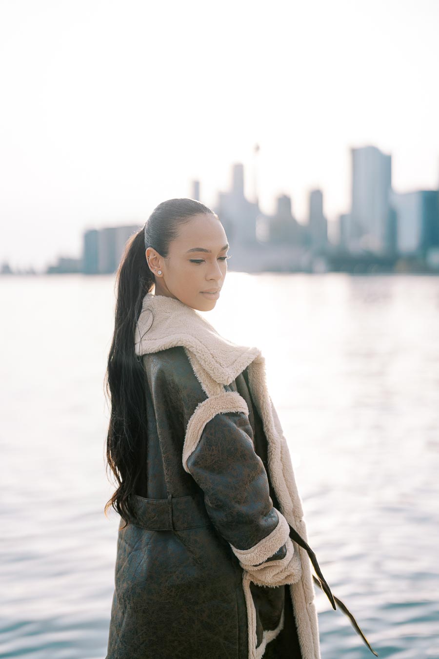 A person in a shearling coat stands by the water with a city skyline in the background, capturing a serene moment.