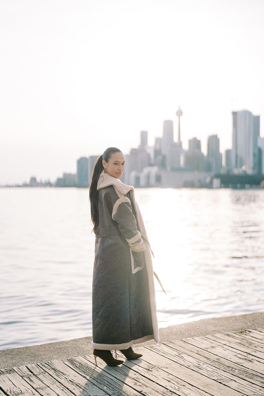 A person stands on a boardwalk by the water, with the Toronto skyline in the background.