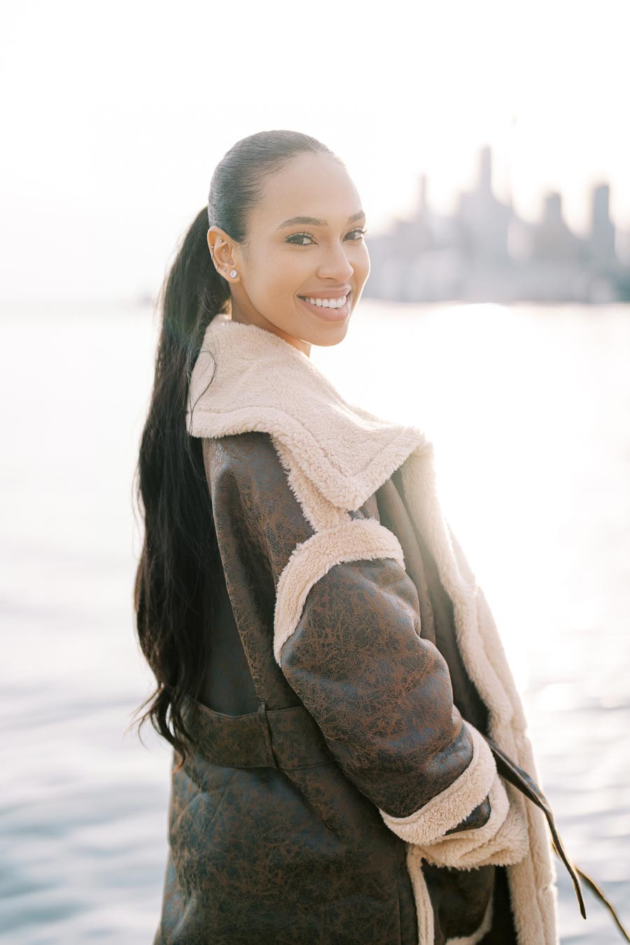 A woman in a brown shearling coat smiles by the water, with a city skyline in the background.