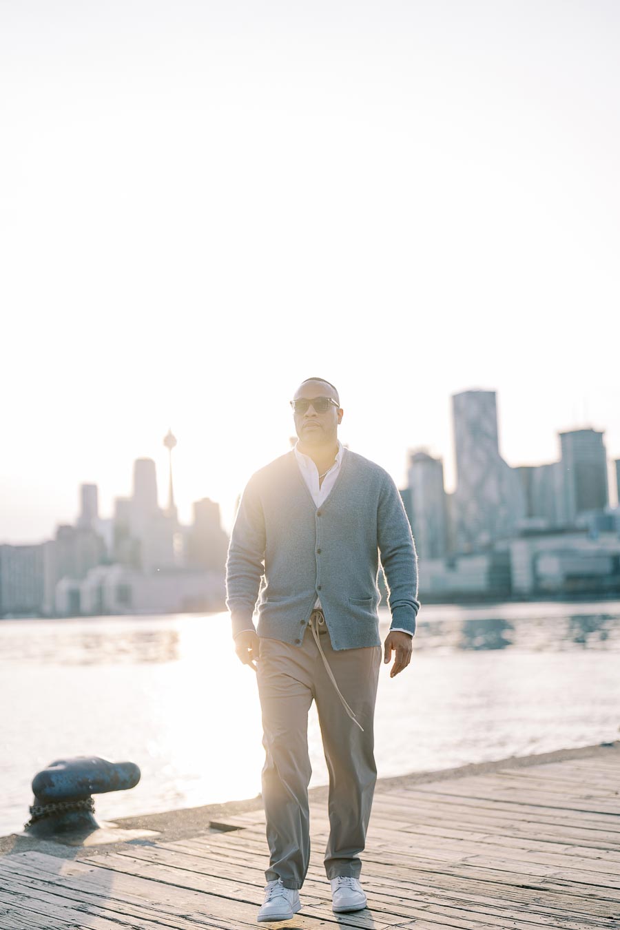 A man walks on a dock with a city skyline in the background, wearing sunglasses and casual attire.
