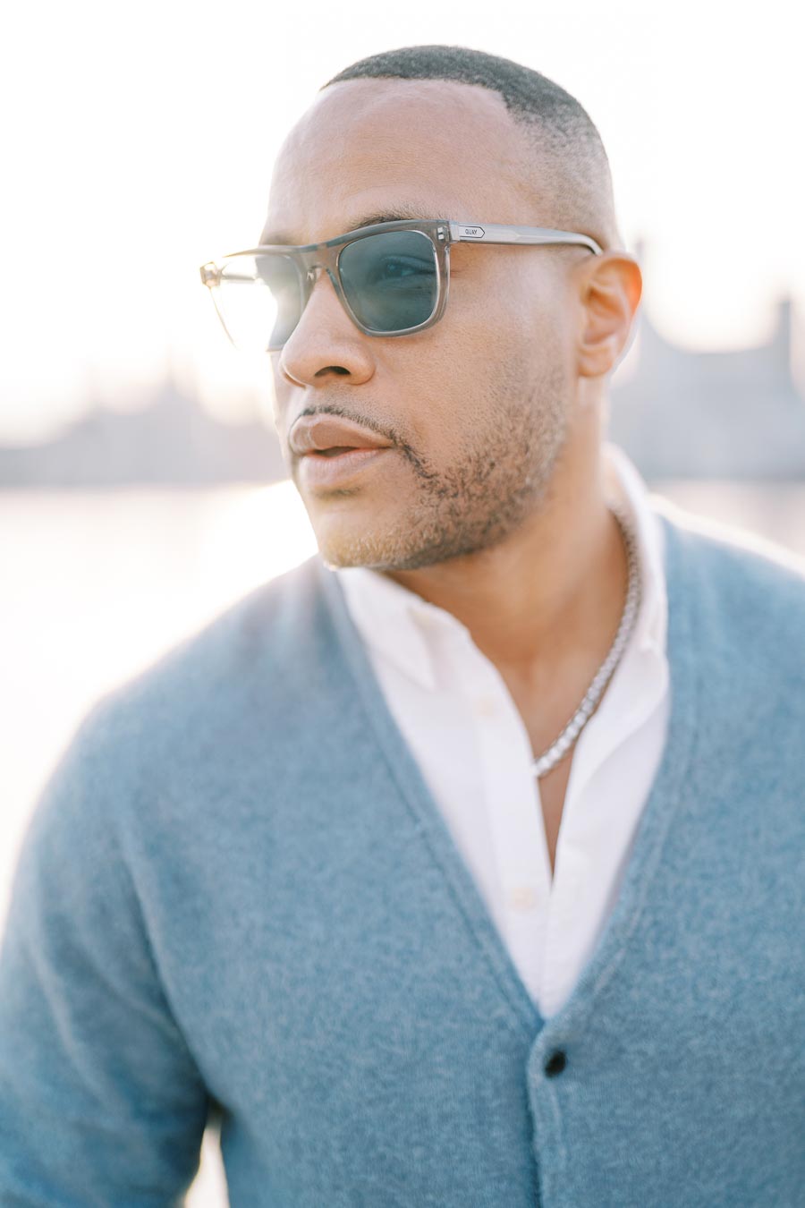 A man in sunglasses and a blue sweater gazes into the distance with a cityscape in the background.