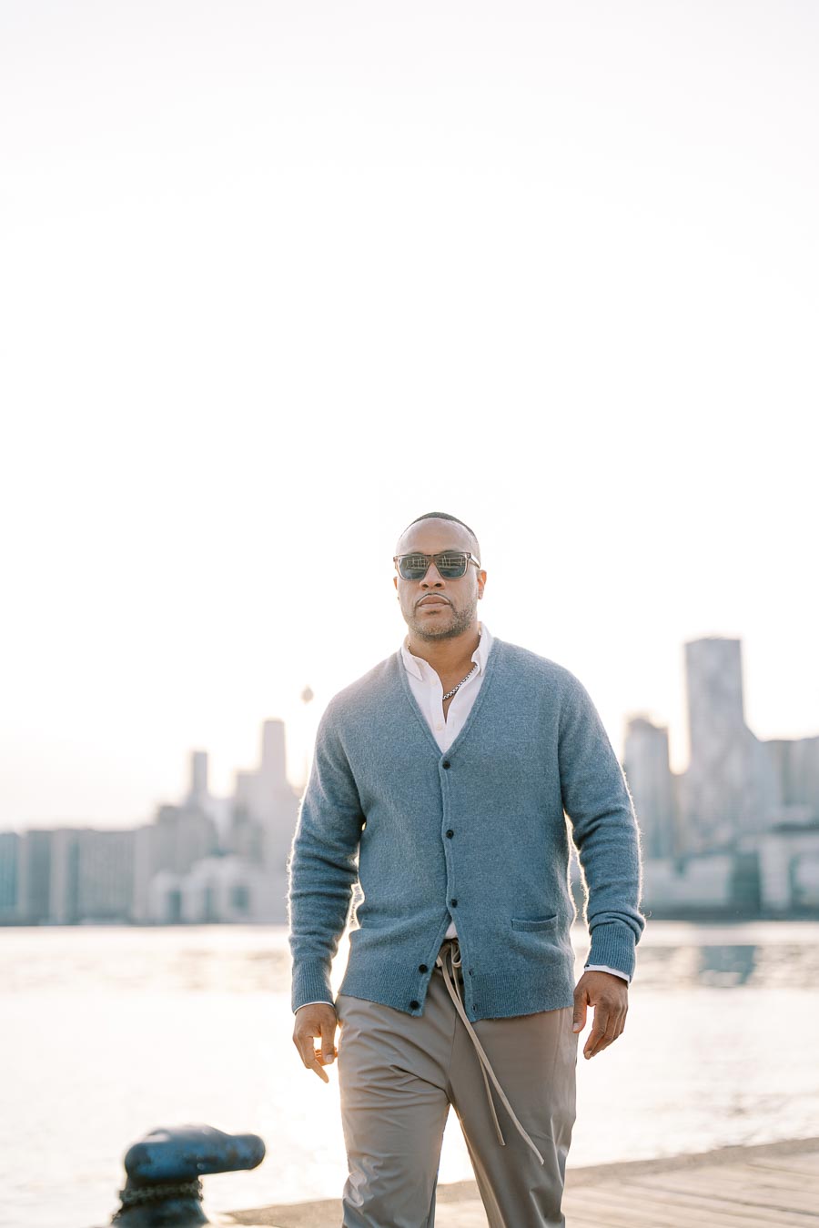 A man in sunglasses and a blue cardigan walks along a waterfront with a city skyline in the background, under a bright sky.