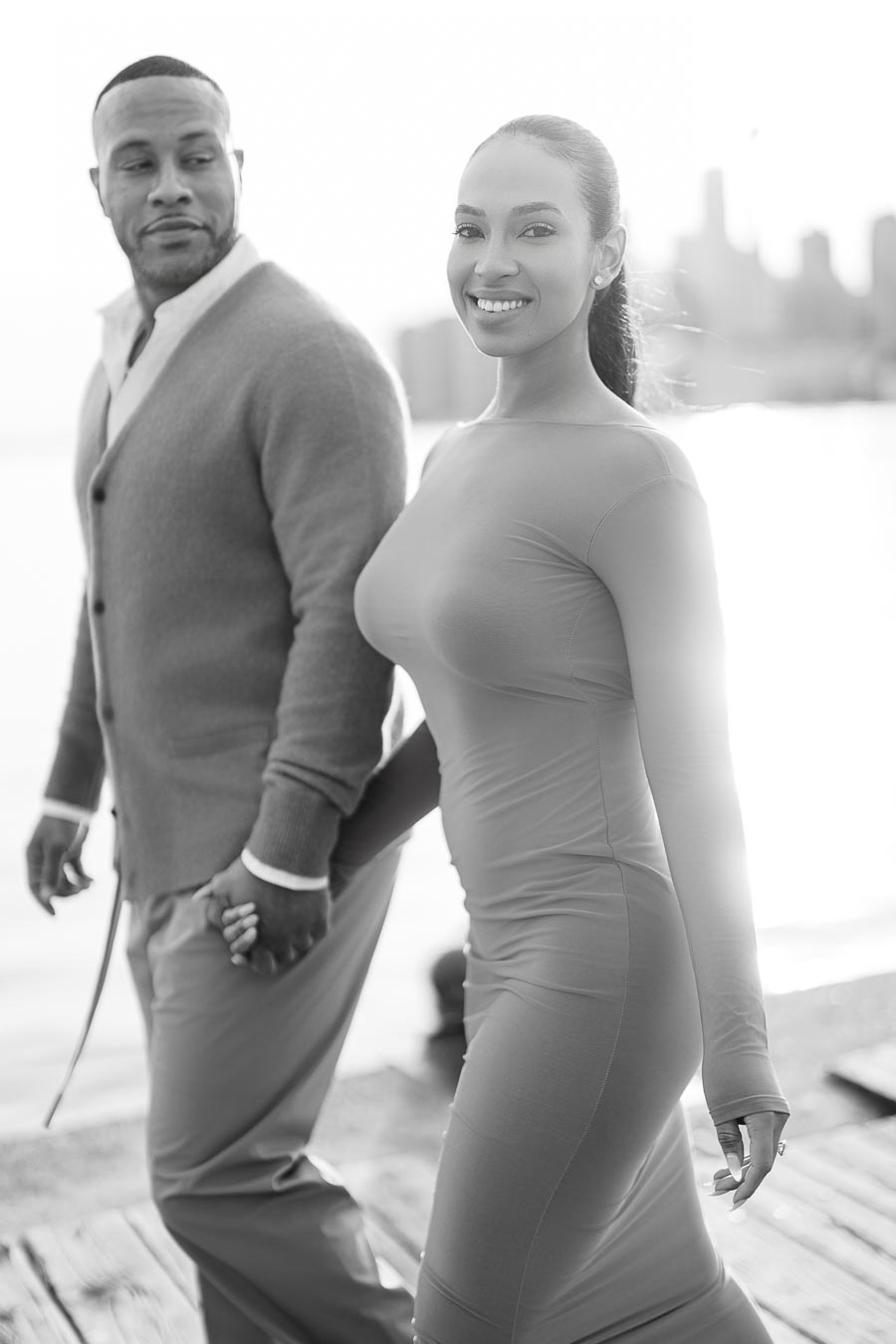 A couple walks hand in hand along a waterfront, with a city skyline in the background, captured in black and white.