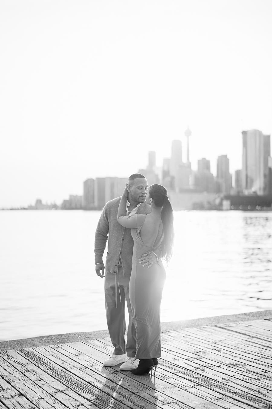 A couple shares a tender moment on a dock with a city skyline in the background, captured in black and white.