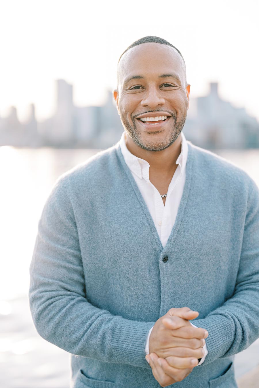 A man in a light blue sweater smiles warmly with a blurred city skyline in the background.