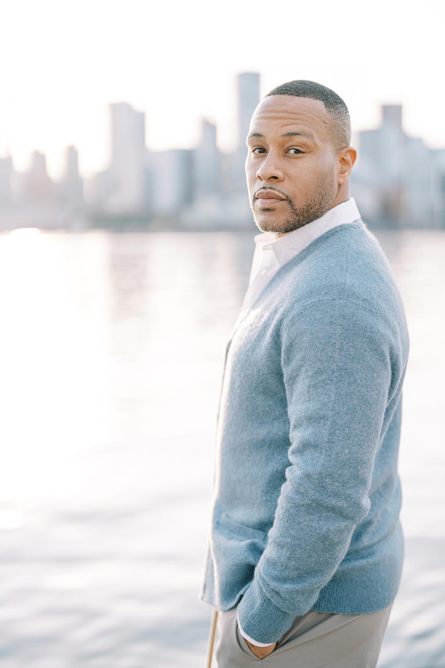 Groom standing by the water in Toronto with hands in pockets, soft sunlight and skyline in background