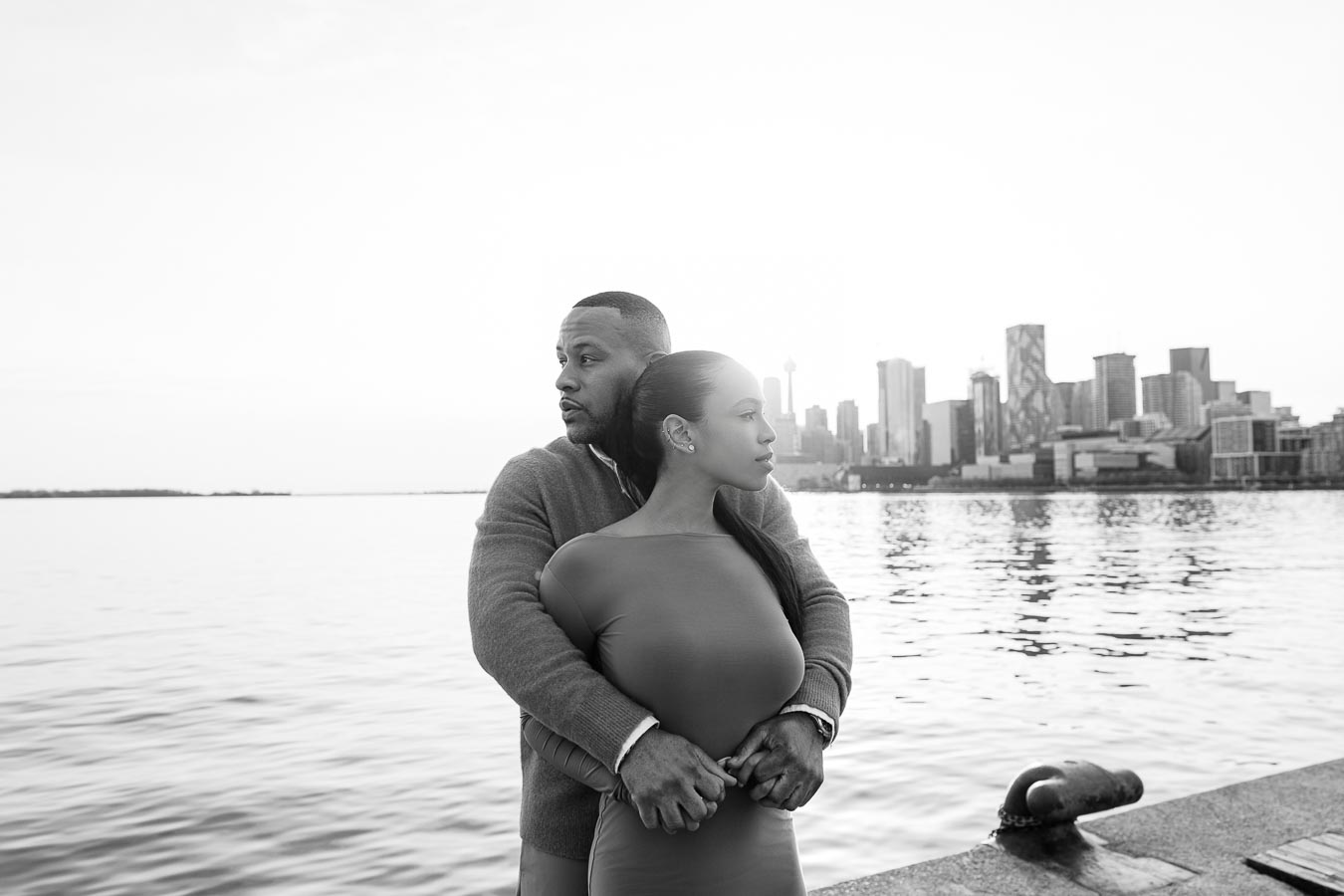 Couple standing together in a black and white portrait at Polson Pier, with the Toronto skyline in the background