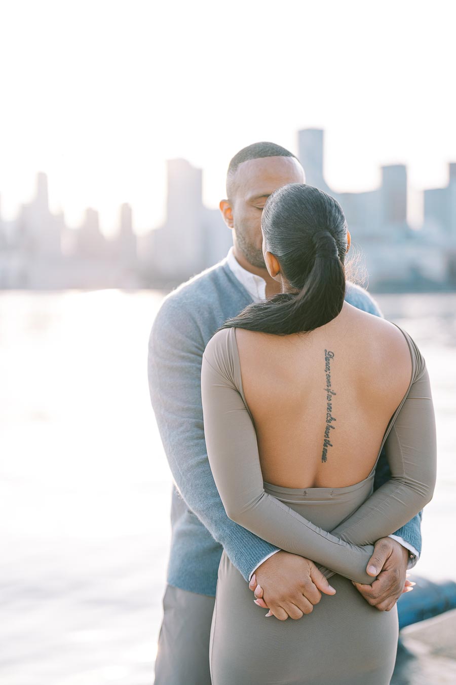 A couple embraces by the water with a city skyline in the background, capturing a romantic moment.