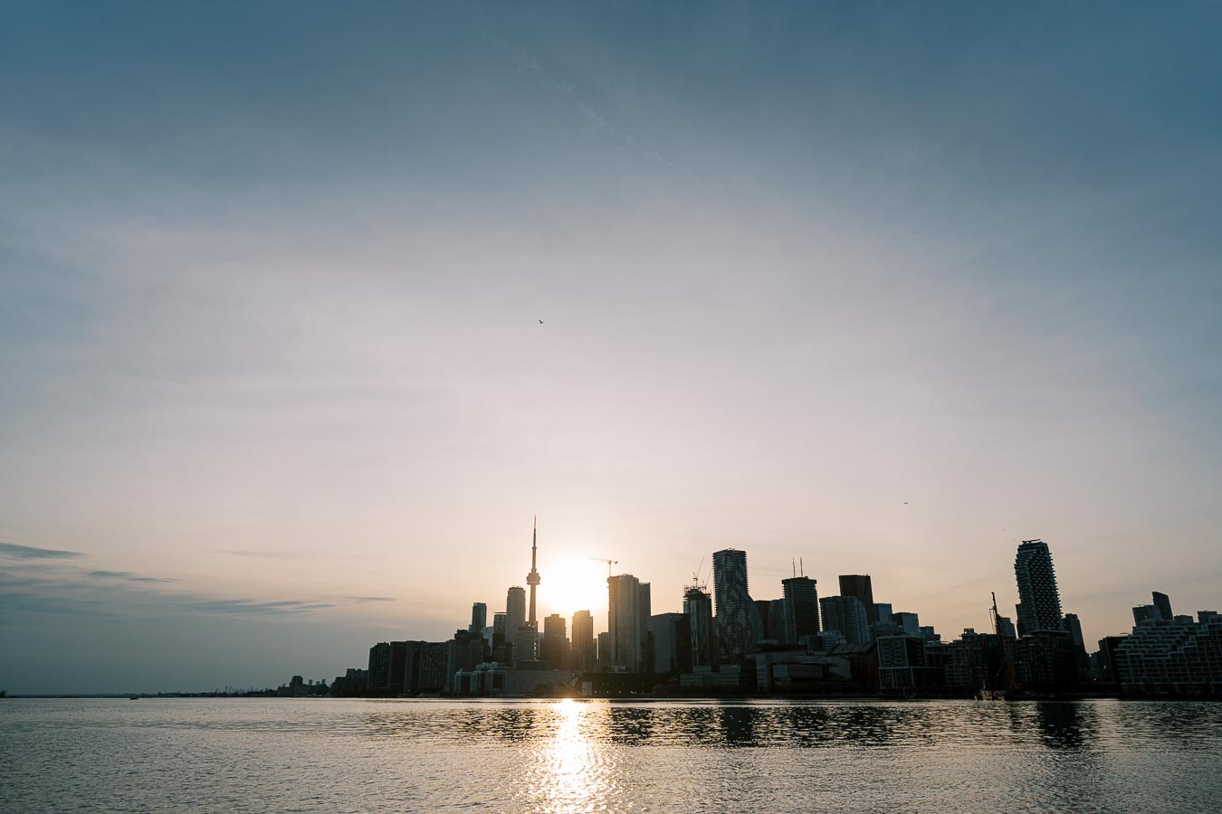 The Toronto skyline is silhouetted against a sunset, with the iconic CN Tower prominently visible. The scene is viewed from across the water, creating a serene and picturesque cityscape.