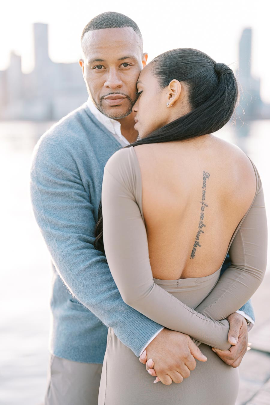 A couple shares an intimate embrace with a city skyline in the background, highlighting the woman's back tattoo and elegant backless dress.