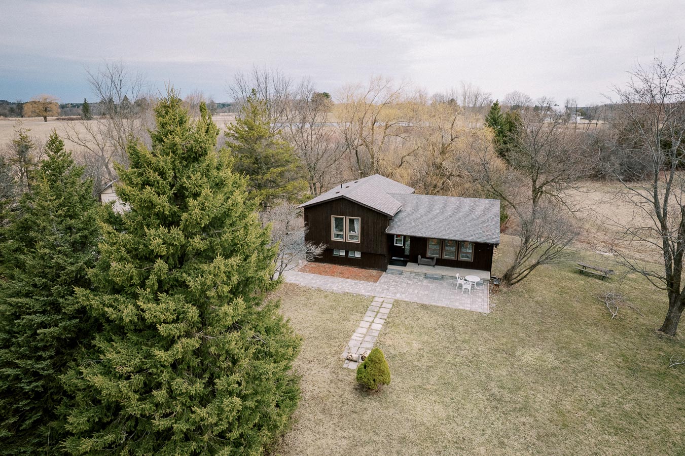 Aerial view of a rustic house surrounded by lush greenery and trees, with a spacious backyard patio featuring outdoor furniture, set in a serene rural landscape under an overcast sky.