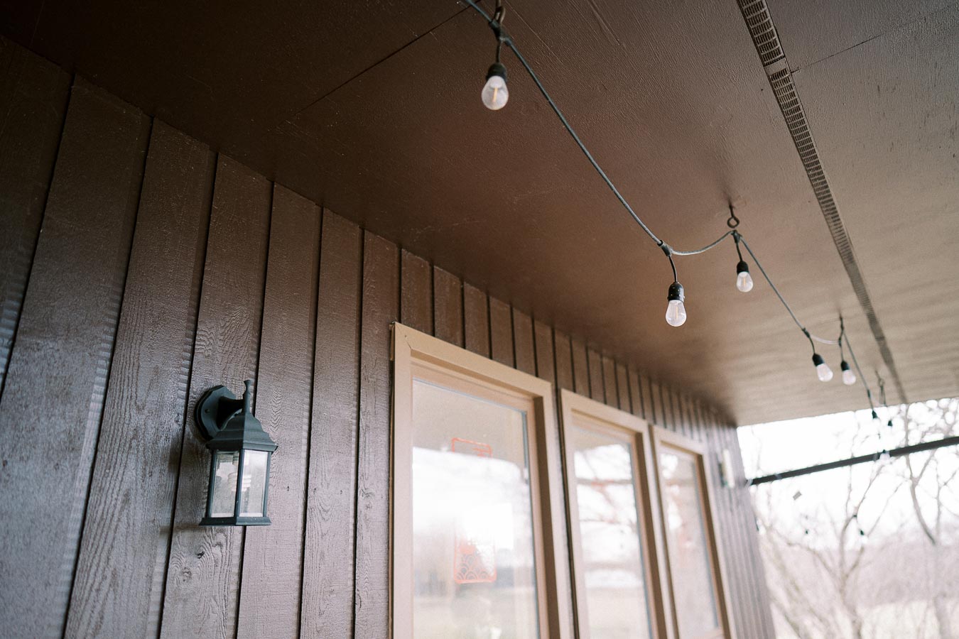 Outdoor patio with string lights hanging from a wooden ceiling above a dark wooden wall with windows.