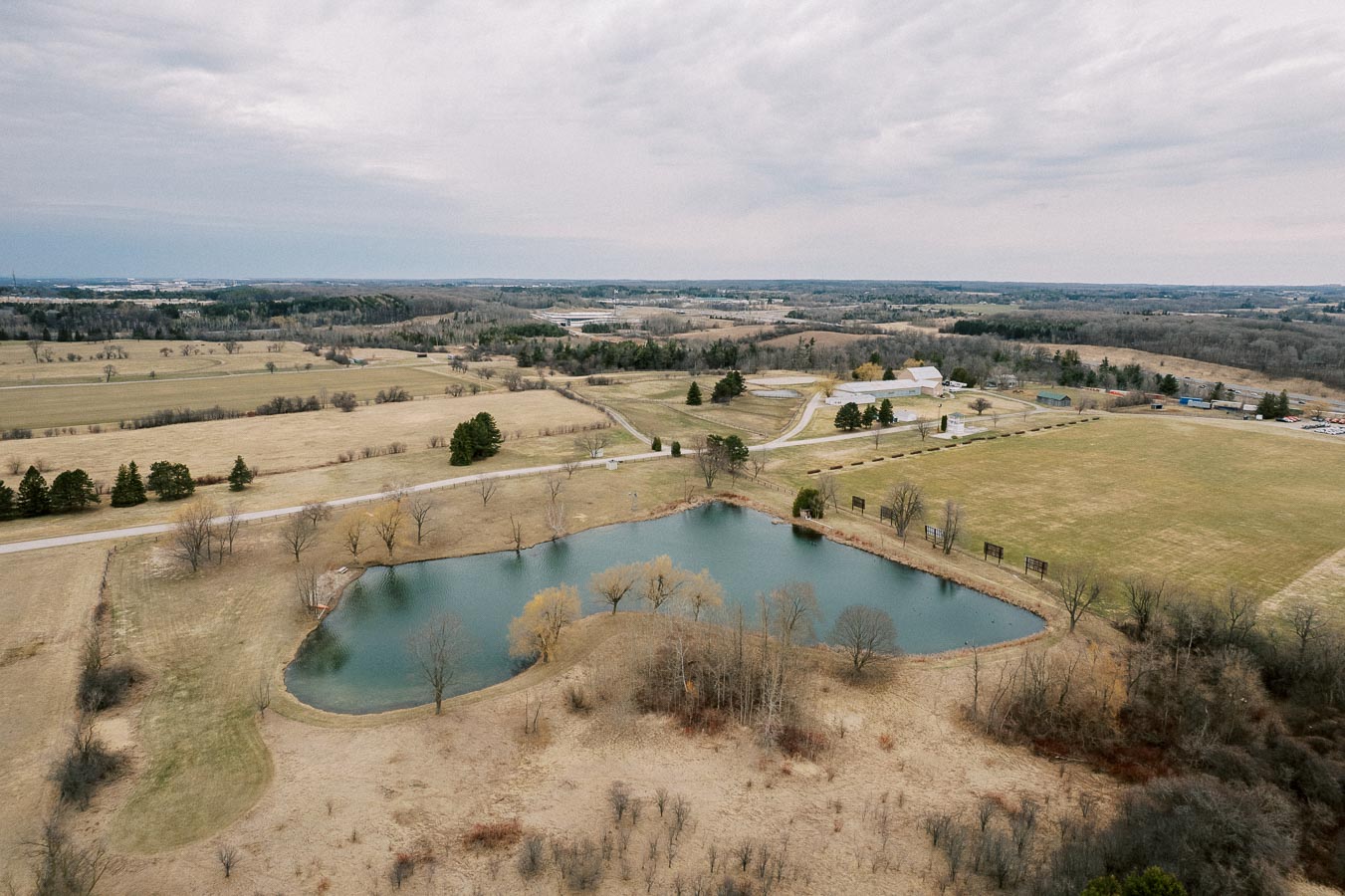 Aerial view of a rural landscape featuring a tranquil pond surrounded by open fields and sparse trees, with farm buildings and rolling hills in the distance under a cloudy sky.