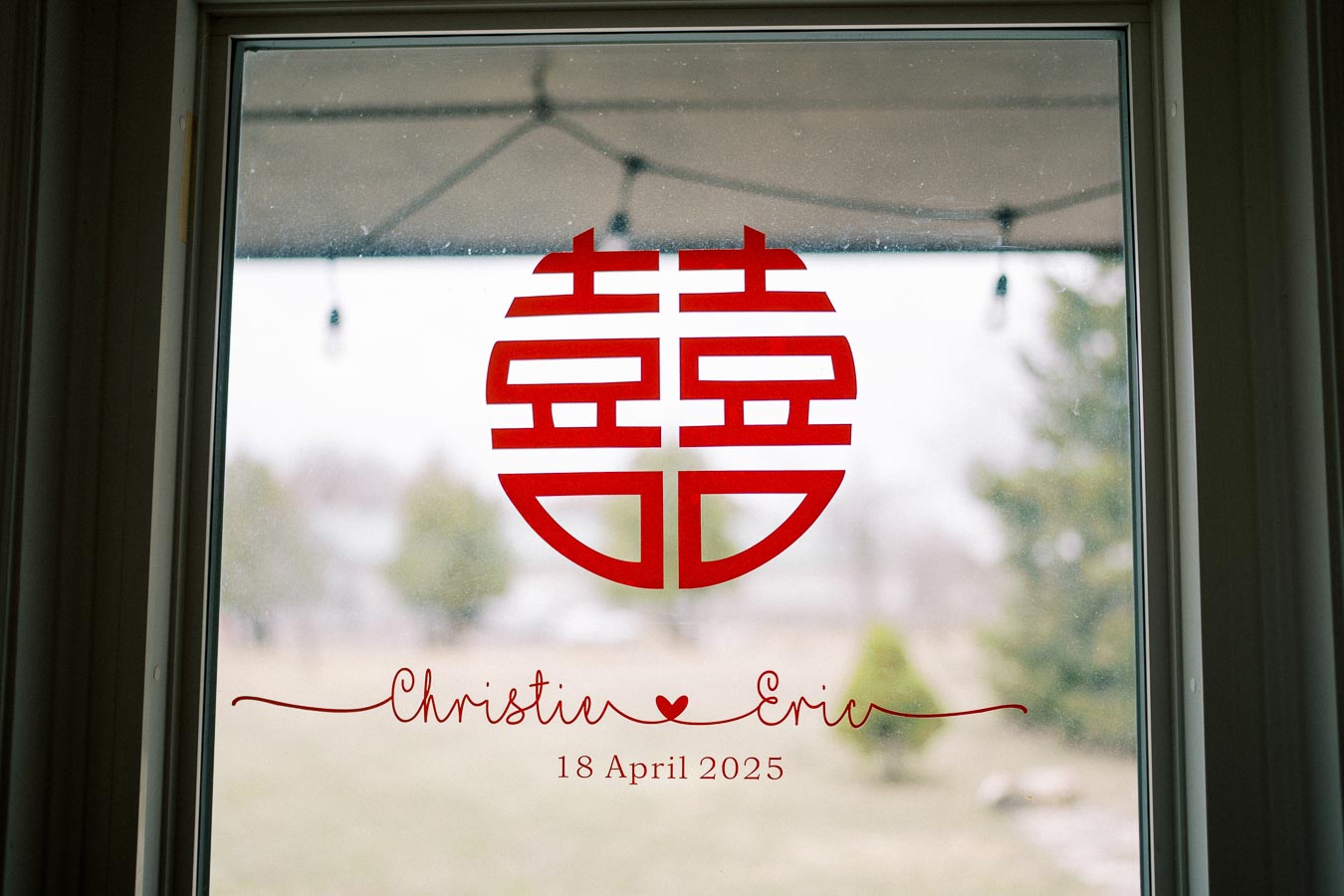 Window decorated with a red Chinese double happiness symbol above the words 'Christie ❤️ Eric, 18 April 2025', symbolizing a wedding celebration.