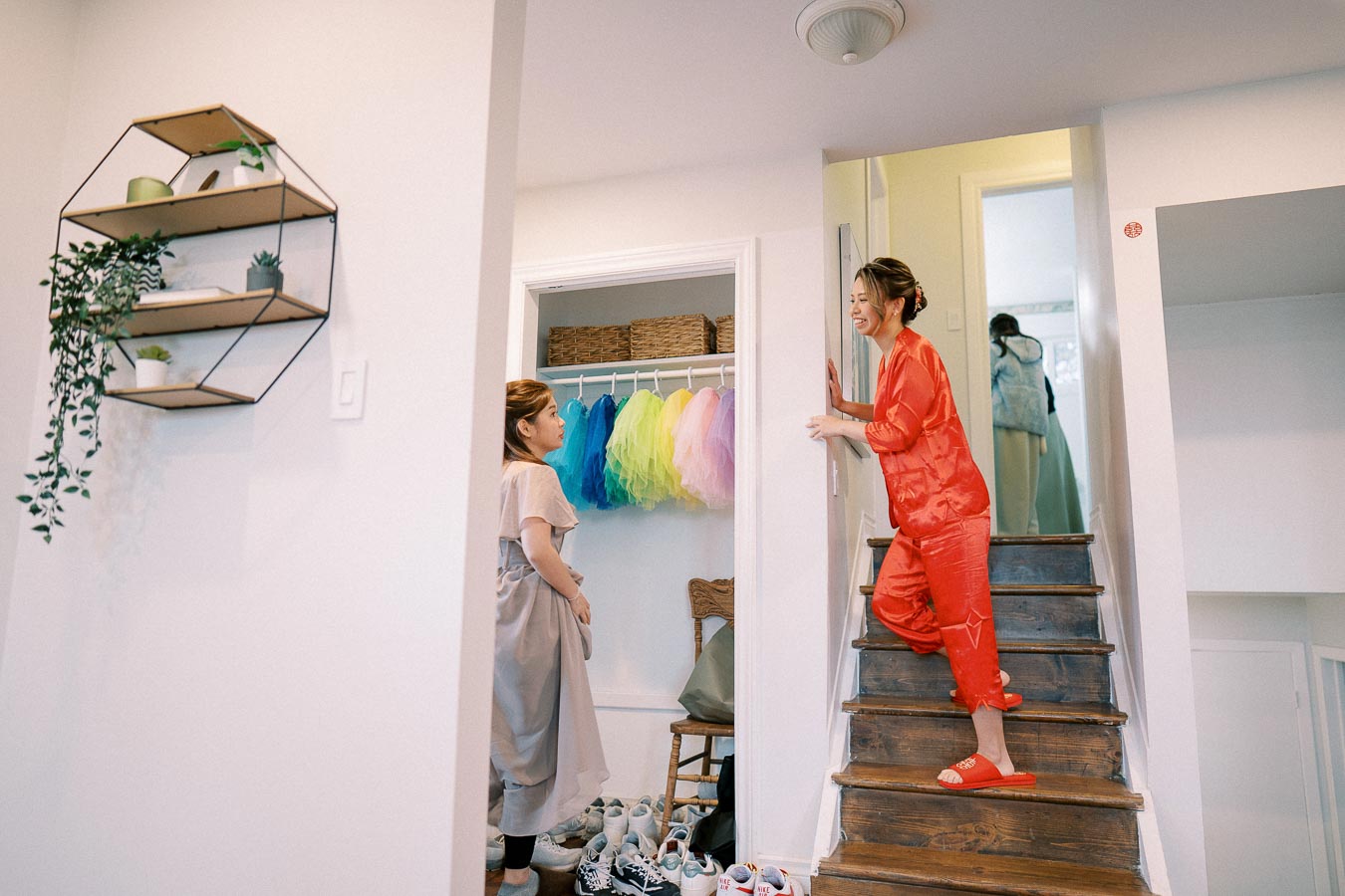 Two women engaged in conversation in a cozy home setting, one standing in a staircase wearing red, with a colorful display of dresses in a closet nearby, alongside shoes neatly arranged. Modern interior design elements include a wall-mounted shelf with plants.