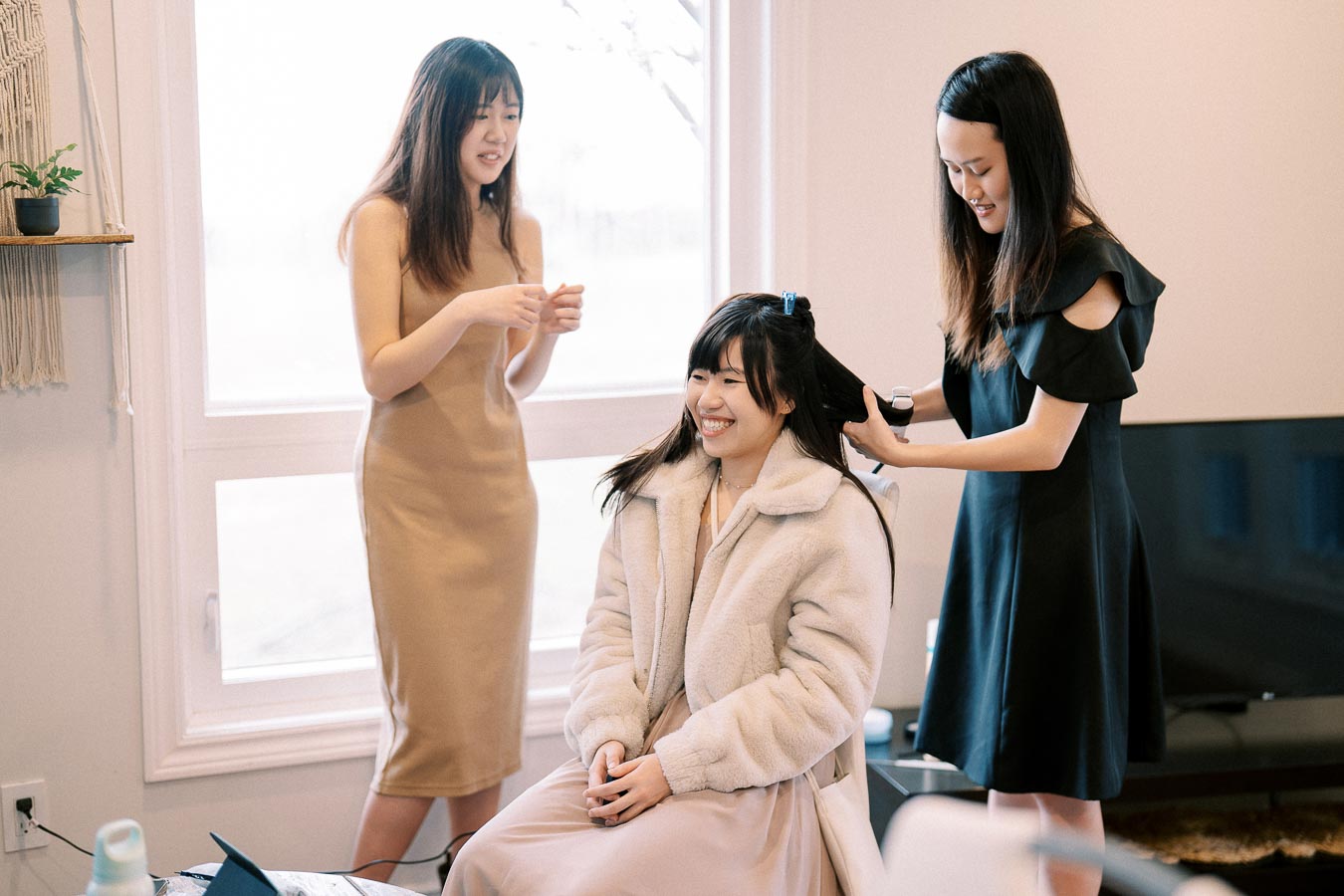 Three women in a bright room, one sitting in a fluffy coat smiling as another styles her hair and the third stands nearby, smiling. Perfect for a lifestyle or fashion context.