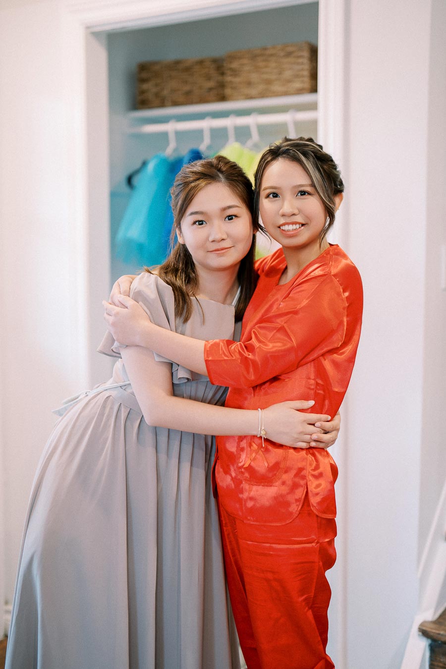 Two women hugging and smiling, dressed in elegant outfits; one in a red silk ensemble, the other in a light gray dress, standing in a room with a closet in the background displaying colorful clothing.
