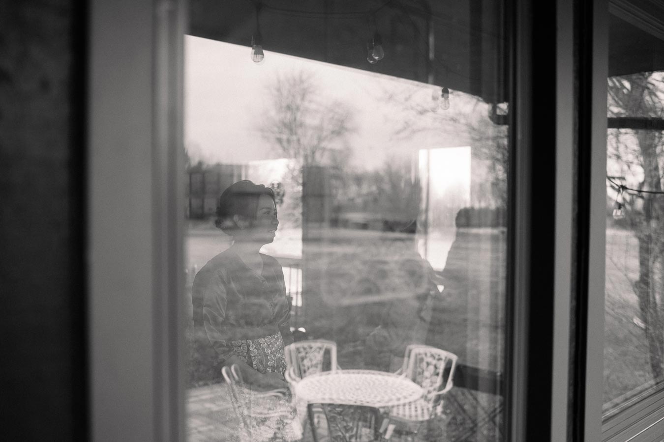 Black and white photo showing a woman reflected in a window, standing near a glass door with a patio table and chairs visible outside. The scene captures a quiet, introspective moment with bare trees in the background.