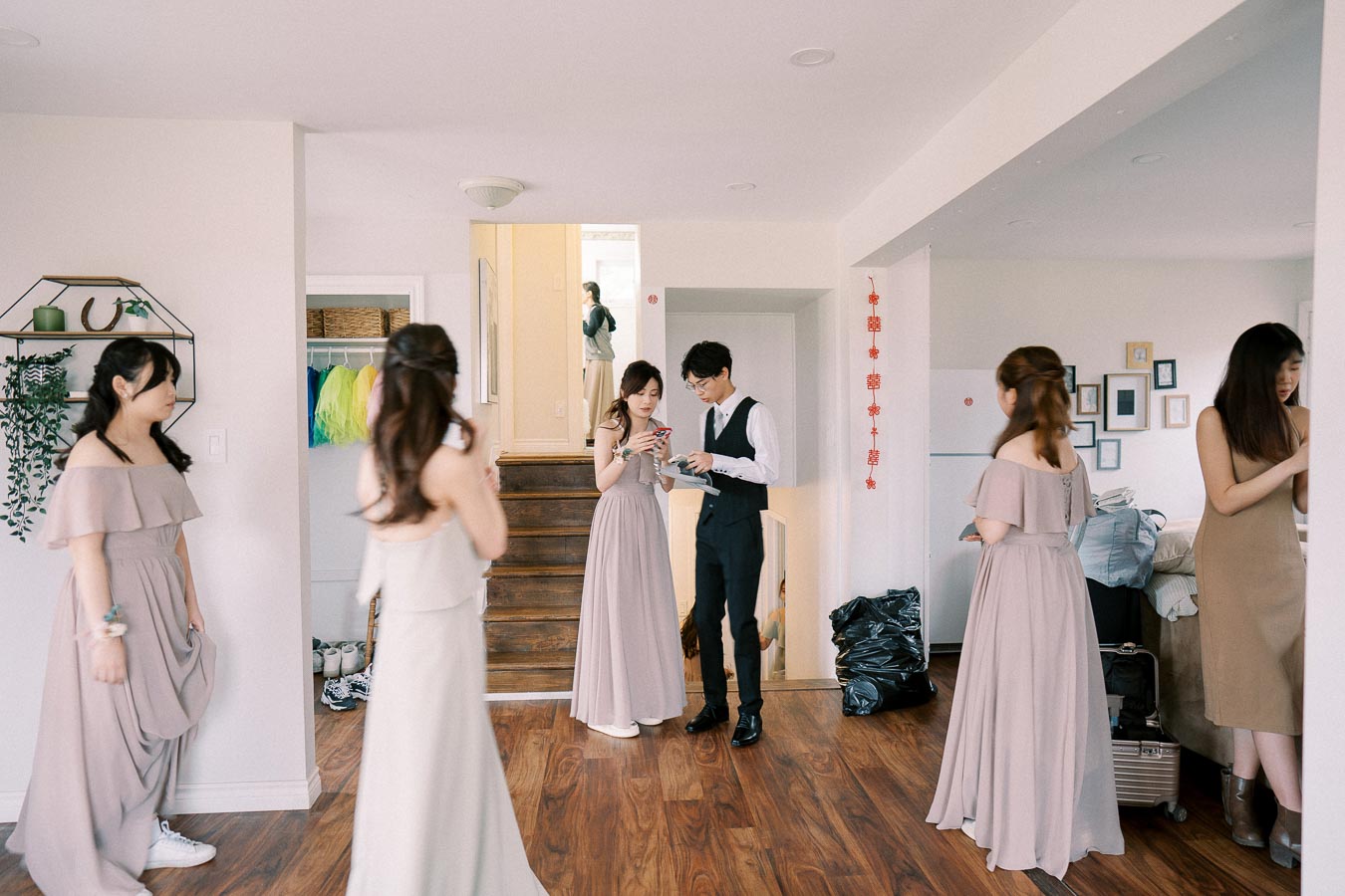 Group of people preparing for a wedding in a bright room, featuring bridesmaids in lavender dresses and a man in formal attire, with a casual atmosphere and home decor.